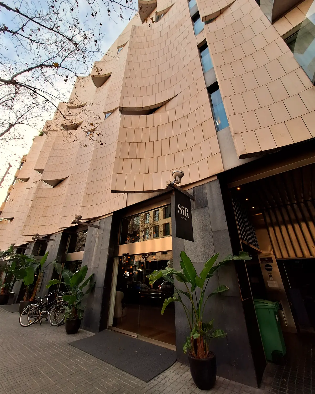 Undulating tiled facade of Sir Victor, with the hotel sign over the glass entrance and planter pots either side