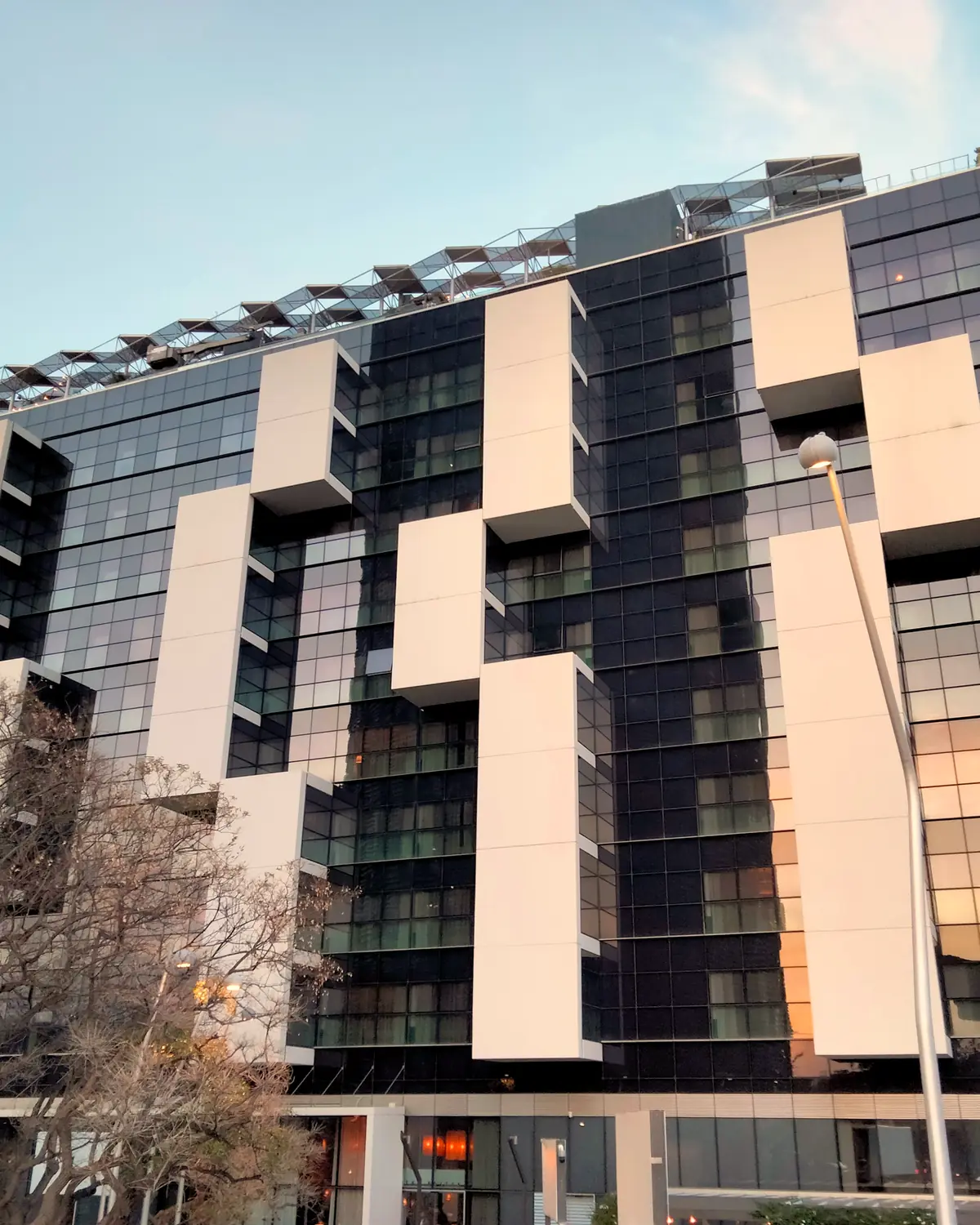 Glass and white-panel facade of The Hoxton, Poblenou at dusk, with a rooftop terrace against a blue sky