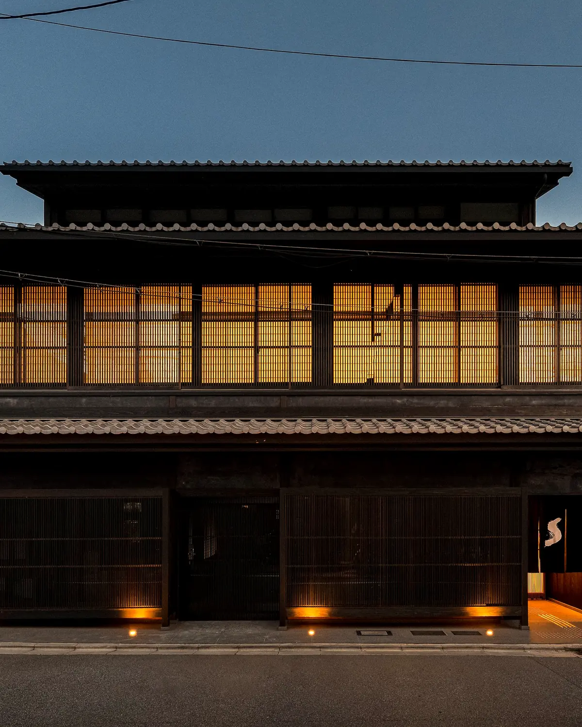 The street facade on Shinmonzen-dori at dusk: a two-storey timber building with tiled roofs, lit lattice windows and a noren at the entrance