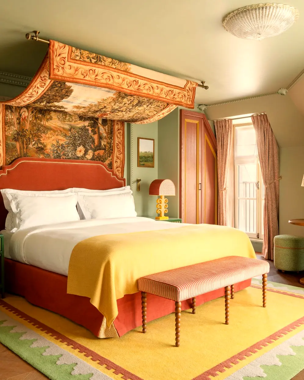 A guest room with a tapestry canopy over a terracotta headboard, a yellow rug and a window onto the street (from the Maisons Pariente press kit)