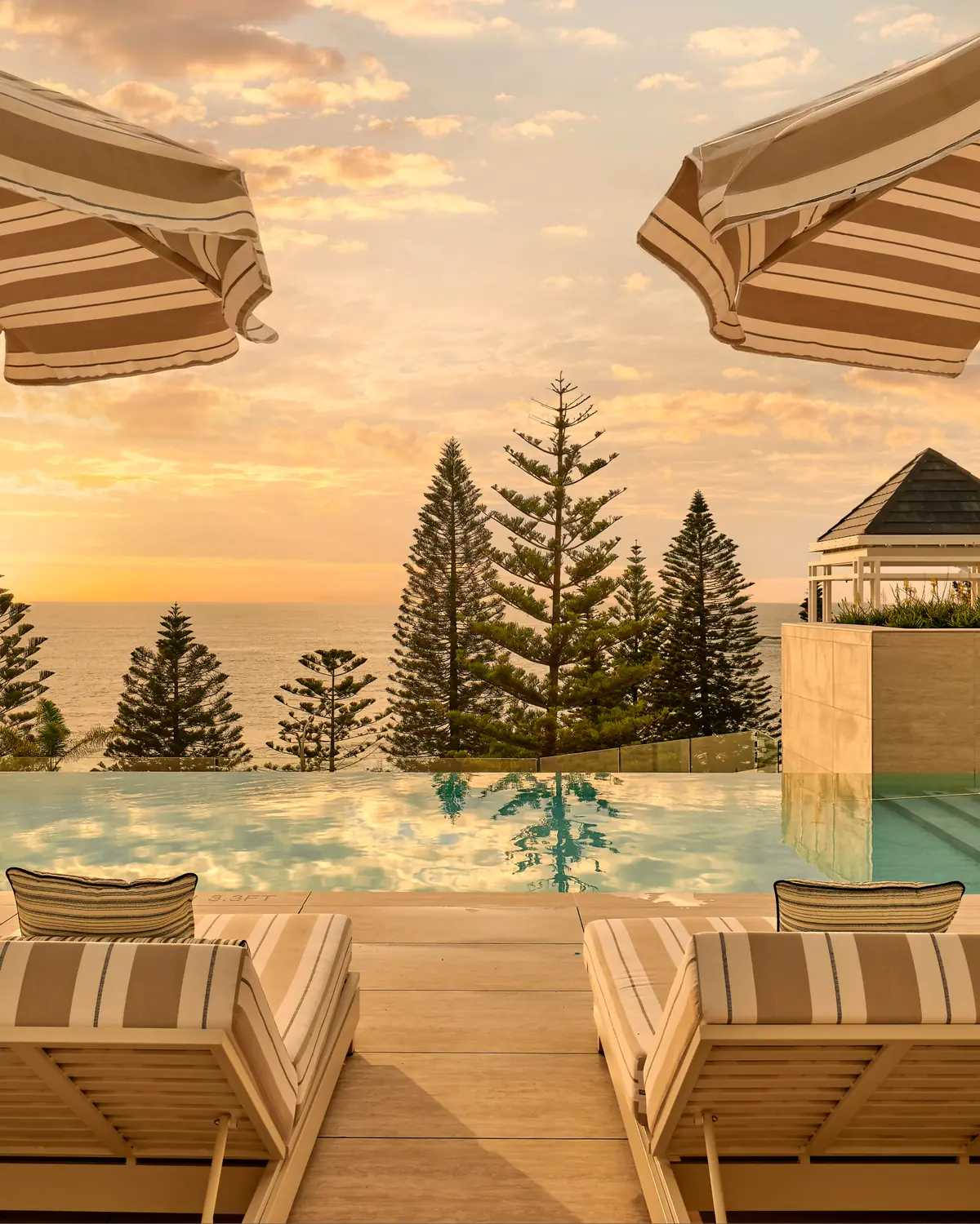 Pool terrace with sun loungers and umbrellas, infinity-edge pool and Norfolk Island pines at sunset (page alt text: "Reimagined interiors and beachfront...