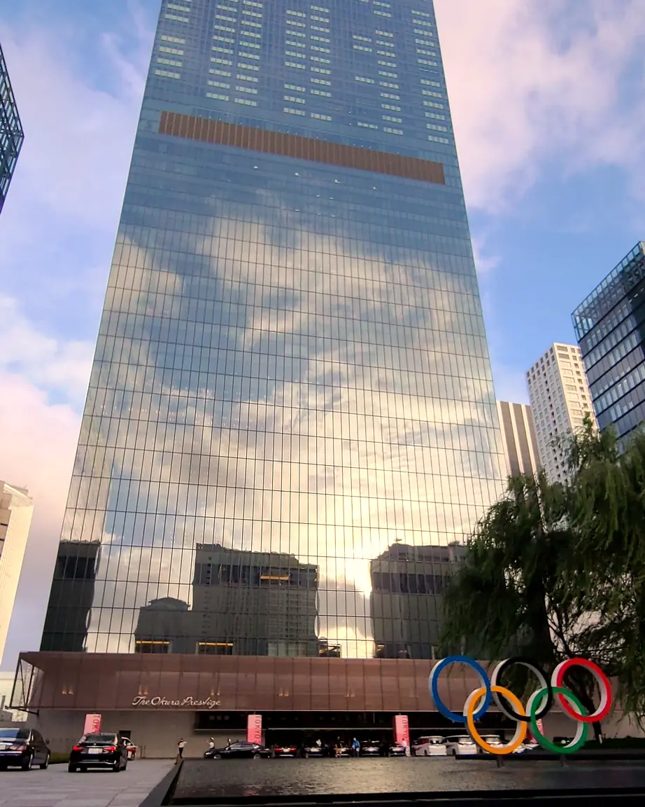 The Okura Prestige Tower (glass tower lettered 'The Okura' at the top and 'The Okura Prestige' over the entrance), ultra-wide view from the forecourt with...